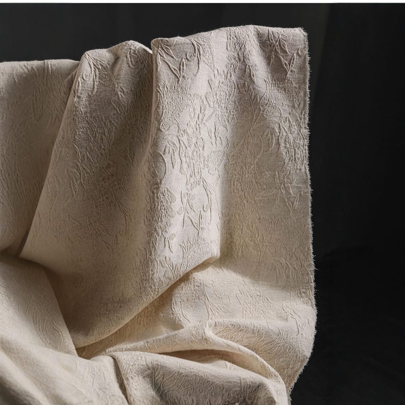May include: Close-up of a cream-colored fabric with a raised floral pattern. The fabric is draped, showing folds and texture. The lighting highlights the intricate design and the soft, neutral color of the material.