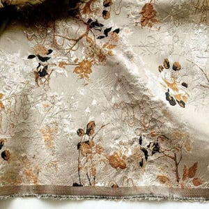 May include: Close-up of a fabric with a floral pattern. The fabric has a light beige background with gold and black floral and branch designs. The fabric has a textured appearance and a frayed edge at the bottom.