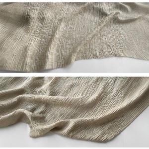Puede incluir: Primer plano de una tela beige con una apariencia texturizada y arrugada. La tela est&aacute; drapeada, con pliegues visibles y sutiles variaciones de tono, creando una sensaci&oacute;n de profundidad y movimiento. El material parece suave y ligero.