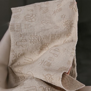 May include: A beige linen fabric with embossed, stylized Asian characters and symbols. The fabric is held up, showing its texture and the detailed design. The overall aesthetic is natural and earthy.