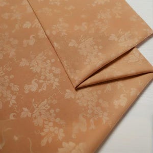 May include: Peach-colored fabric with a repeating pattern of grape vines and swan-like figures. The fabric is folded, showcasing the detailed design and texture. Ideal for home decor or crafting projects.