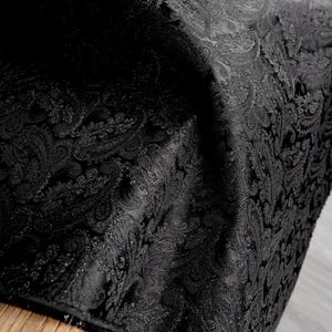 Może przedstawiać: Zbliżenie czarnej tkaniny w paisley. Tkanina ma szczegółowy, powtarzalny wzór o lekko teksturowanym wyglądzie. Ciemny kolor i skomplikowany wzór sugerują luksusowy materiał, prawdopodobnie do tapicerki lub odzieży.