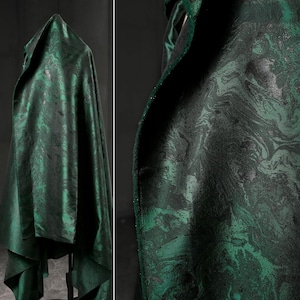 Puede incluir: Una tela verde oscuro con un patrón veteado, drapeada sobre un soporte. La tela presenta tonos de verde y negro, creando un efecto arremolinado. El primer plano revela los intrincados detalles del diseño y su textura.