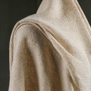 May include: Close-up of a beige fabric draped over a shoulder. The fabric has a textured weave and a natural, neutral color. The folds create shadows and depth, highlighting the material's texture.