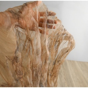 May include: Close-up of sheer, flowing fabric in shades of tan and brown. The delicate material is held up, revealing its texture and the light passing through it. The fabric has a crinkled appearance.