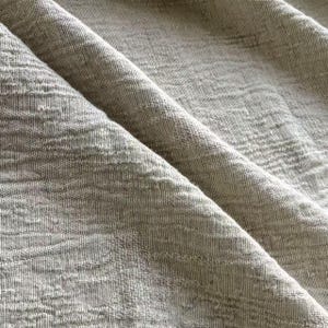 May include: Close-up of a folded, textured fabric in a neutral, light gray color. The fabric has a subtle, embossed pattern and a soft, natural appearance. The folds create shadows and depth, highlighting the texture.