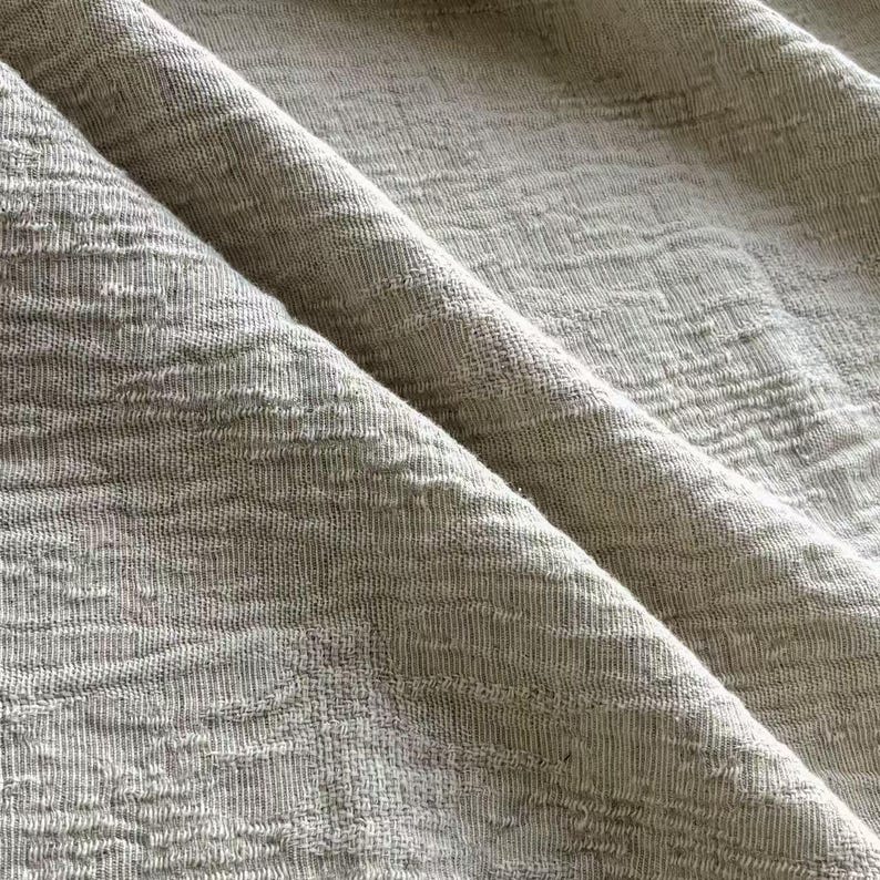 May include: Close-up of a folded, textured fabric in a neutral, light gray color. The fabric has a subtle, embossed pattern and a soft, natural appearance. The folds create shadows and depth, highlighting the texture.