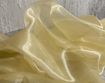 May include: Close-up of a flowing, sheer fabric in a soft, golden-yellow hue. The fabric has a subtle sheen, with folds and curves that catch the light, creating highlights and shadows. The material appears delicate and lightweight.