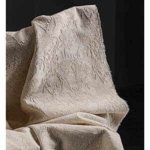 May include: A close-up of a beige fabric with a raised floral pattern. The fabric is draped, showing the texture and design. The neutral color and detailed pattern suggest a decorative textile, possibly for home decor or apparel.