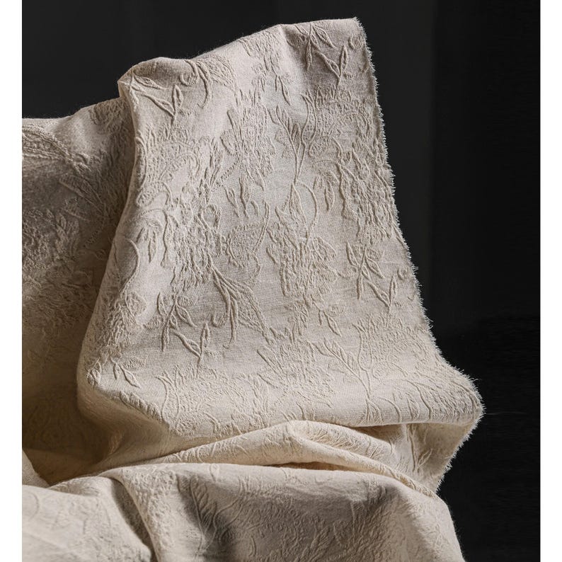 May include: A close-up of a beige fabric with a raised floral pattern. The fabric is draped, showing the texture and design. The neutral color and detailed pattern suggest a decorative textile, possibly for home decor or apparel.