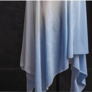 May include: A flowing fabric with a gradient of colors, transitioning from white to light blue. The fabric is draped, creating soft folds and a layered effect. The overall aesthetic is airy and elegant.