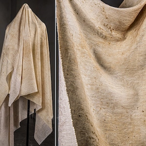 May include: A draped, off-white fabric with a textured surface and subtle brown speckles. The fabric is hanging on a black stand, with a close-up view showing intricate details of the weave and pattern.