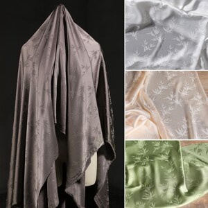 May include: A draped fabric in a dark gray color with a subtle bamboo leaf pattern. Additional fabric swatches in white, cream, and green are also shown. The fabric has a silky sheen, suggesting it is made of silk or a similar material.