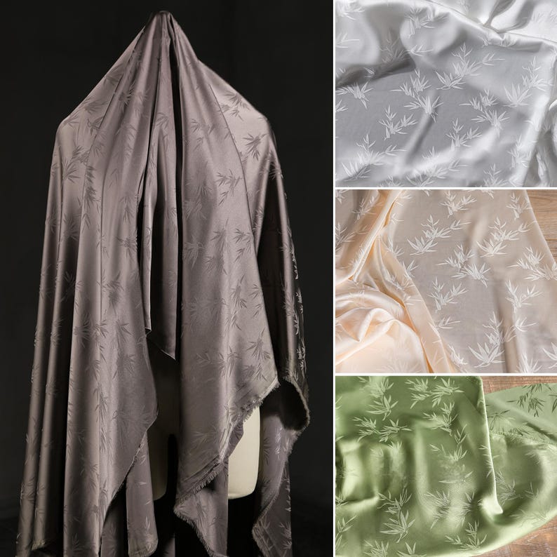May include: A draped fabric in a dark gray color with a subtle bamboo leaf pattern. Additional fabric swatches in white, cream, and green are also shown. The fabric has a silky sheen, suggesting it is made of silk or a similar material.