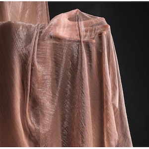 Peut inclure: Un tissu transparent et chatoyant de couleur rose pâle. Le tissu est drapé, avec des lignes diagonales visibles et un éclat subtil. Le matériau semble léger et fluide, la lumière le traversant. Le fond est sombre.