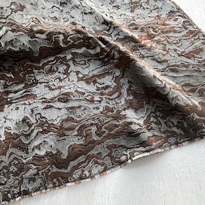 May include: A close-up of a fabric with a marbled pattern. The fabric features a blend of gray and brown tones, creating a textured, flowing design. The material appears to be soft and draped, with visible folds and subtle variations in color.