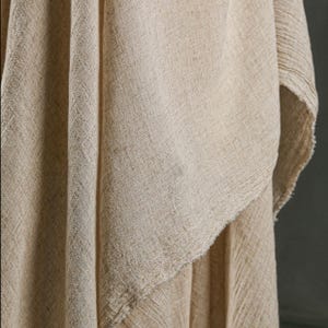 May include: Close-up of a beige linen fabric with a textured weave. The fabric is draped, showing folds and a raw, unfinished edge. The neutral color and natural texture suggest a focus on home decor or textile crafts.