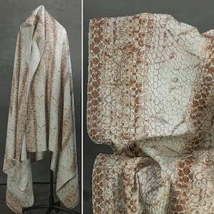 Puede incluir: Un textil drapeado con un estampado de piel de serpiente en tonos beige y marrón. La tela se exhibe sobre un soporte negro, resaltando su textura y diseño. El primer plano revela el detallado patrón de escamas.