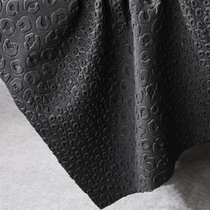 Peut inclure: Gros plan sur un tissu noir avec un motif léopard en relief et texturé. Le tissu est plié, révélant le design et les ombres complexes. Le matériau semble doux et souple.