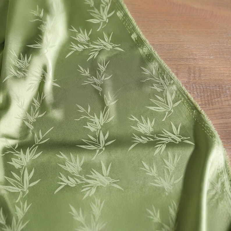 May include: Close-up of a green fabric with a bamboo leaf pattern. The fabric has a satin sheen and a light green color. The bamboo leaves are a lighter shade of green, creating a subtle contrast. The fabric is folded, showing its texture.