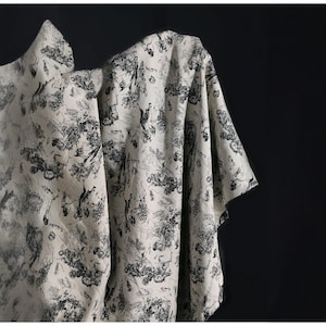 May include: Off-white fabric with a black toile pattern, draped against a dark background. The fabric features a detailed design of trees, figures, and architectural elements. The fabric is folded, showing the pattern's repeat.