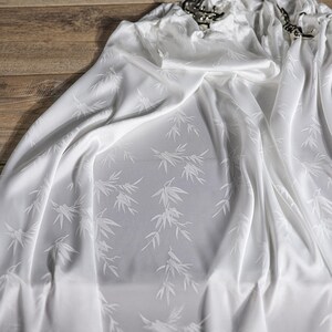 May include: White fabric with a subtle bamboo leaf pattern. The fabric is draped, showing soft folds and a silky texture. Decorative metal curtain rings are visible at the top.