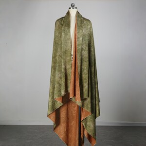 May include: A draped, reversible shawl on a mannequin. The outer layer is olive green with a mottled pattern, while the interior is a warm, rust-colored fabric with a subtle floral design. The shawl is displayed in a studio setting.