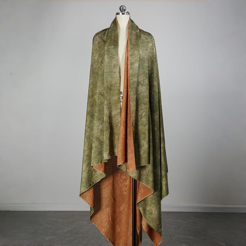 May include: A draped, reversible shawl on a mannequin. The outer layer is olive green with a mottled pattern, while the interior is a warm, rust-colored fabric with a subtle floral design. The shawl is displayed in a studio setting.