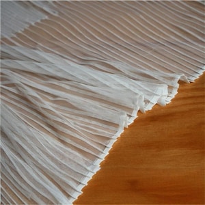 May include: Close-up of white pleated fabric with a delicate, sheer texture. The fabric is arranged in diagonal folds, creating a wave-like pattern. The background is a warm, wooden surface.