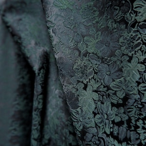May include: Close-up of a dark green floral brocade fabric. The fabric has a textured surface with a repeating pattern of flowers and leaves. The fabric is folded, creating shadows and depth.