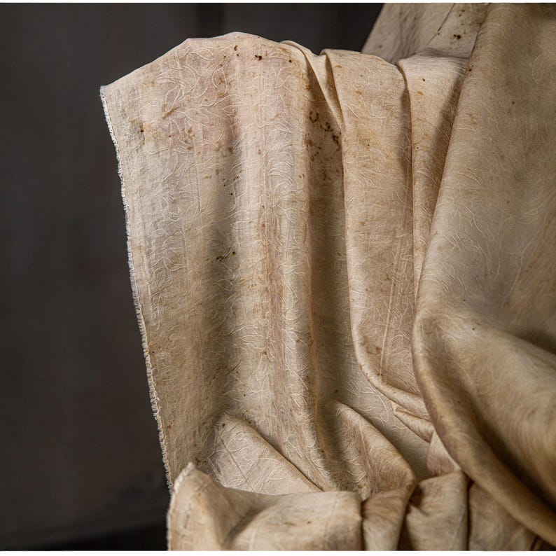 May include: Close-up of a beige fabric with a subtle, textured pattern. The fabric is draped, showing folds and creases. The color is a warm, neutral tone with darker spots, suggesting a natural dye or aging process. The edges are unfinished.