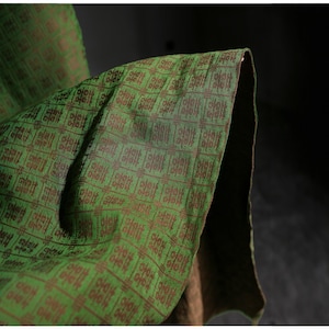 Puede incluir: Primer plano de una tela verde con un patrón geométrico marrón repetido. La tela está doblada, revelando un vistazo de un reverso más oscuro, posiblemente marrón. La textura parece suave y posiblemente tejida.