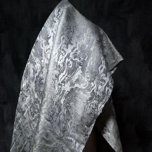 May include: A draped fabric with a marbled pattern in shades of gray and white. The fabric has a flowing, organic design, with swirling lines and textures. The material appears to be soft and luxurious, with a subtle sheen. The background is a dark, solid color.