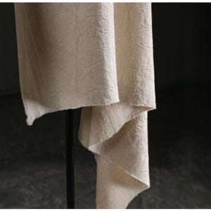 May include: Close-up of a beige fabric with a textured, floral-like pattern. The fabric is draped, revealing its texture and the way it hangs. The edges are unfinished, adding to the natural aesthetic. The fabric is likely used for home decor.