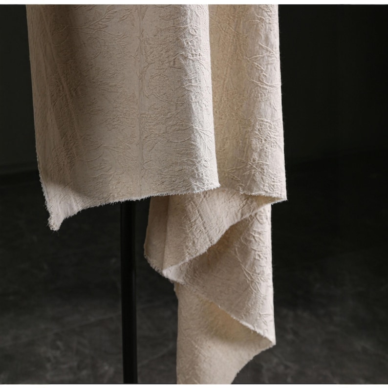 May include: Close-up of a beige fabric with a textured, floral-like pattern. The fabric is draped, revealing its texture and the way it hangs. The edges are unfinished, adding to the natural aesthetic. The fabric is likely used for home decor.