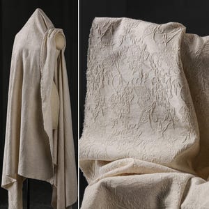 May include: Beige fabric with a raised floral pattern, draped over a mannequin and displayed in a close-up. The fabric has a textured appearance and a raw edge. The floral design is visible in the close-up, showcasing the intricate details of the fabric.