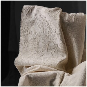 May include: Close-up of a cream-colored fabric with a raised floral pattern. The fabric is folded, revealing the texture and design. The neutral color and detailed pattern suggest a decorative textile.