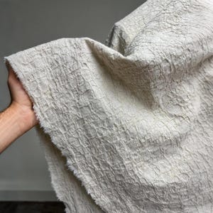 Könnte beinhalten: Nahaufnahme eines strukturierten, cremefarbenen Stoffes mit einem gewebten Muster. Der Stoff hat einen rohen, ausgefransten Rand und wird hochgehalten, wodurch seine Falten und Textur sichtbar werden. Das Material scheint Leinen oder eine ähnliche Naturfaser zu sein.