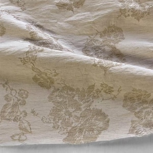 Puede incluir: Primer plano de una tela beige con un estampado floral. El diseño presenta flores y follaje detallados en relieve en un tono beige más claro, creando un contraste sutil. La tela parece ligeramente texturizada y drapeada.
