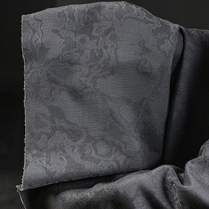 May include: A close-up shot of a dark grey fabric with a delicate floral pattern. The fabric is folded, showing its texture and the intricate design. The material appears soft and drapes elegantly, suitable for clothing or home decor.