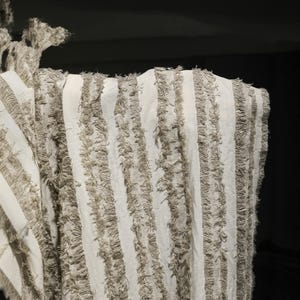 May include: Close-up of a textile with vertical stripes. The fabric is off-white with textured, fringed stripes in a neutral, light brown color. The edges of the fabric are also fringed. The textile appears to be a decorative element.