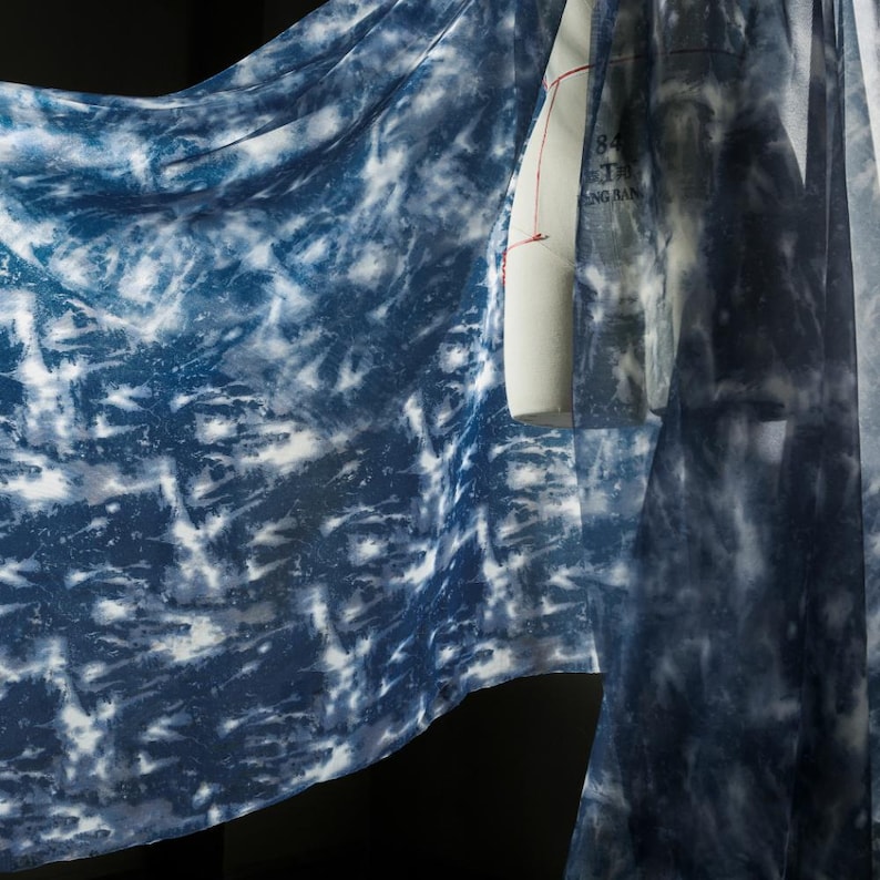 May include: A draped fabric featuring a tie-dye design in blue and white hues. The material is patterned with a marbled effect, with deep indigo sections contrasting with lighter, almost white, areas. A dress form is partially visible.