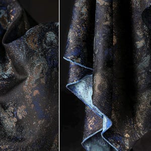 May include: Close-up of a dark fabric with a textured, speckled design. The fabric features a mix of navy blue, teal, and gold tones, creating a shimmering effect. The edges reveal a light blue underside, adding contrast.