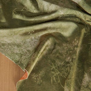 May include: Close-up of a draped, olive green fabric with a subtle floral and foliage pattern. The fabric has a soft sheen and a textured appearance, with folds and shadows adding depth. The underside of the fabric is a contrasting orange color.