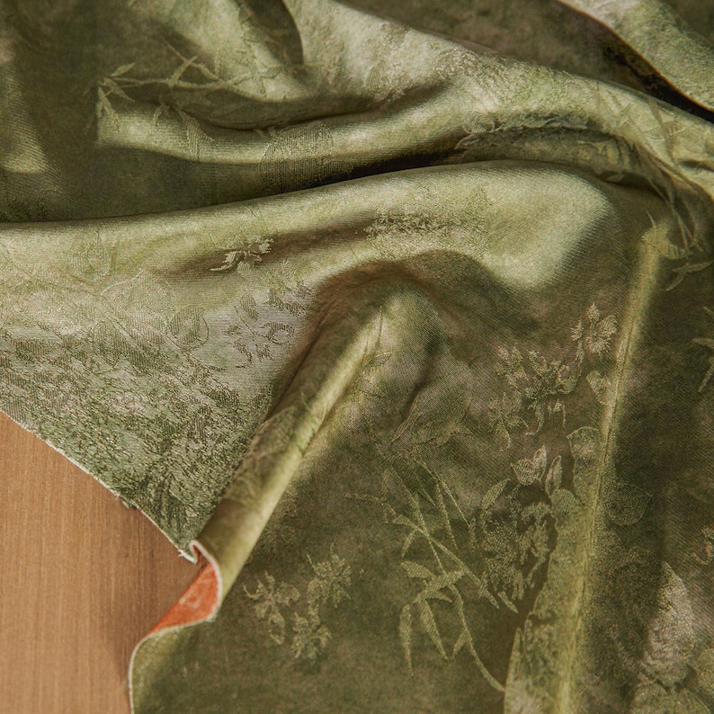 May include: Close-up of a draped, olive green fabric with a subtle floral and foliage pattern. The fabric has a soft sheen and a textured appearance, with folds and shadows adding depth. The underside of the fabric is a contrasting orange color.