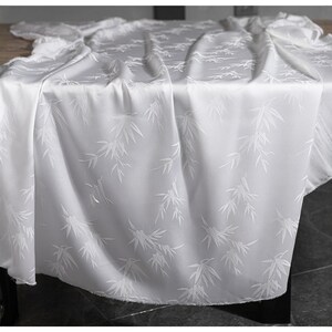 May include: A white tablecloth with a subtle bamboo leaf pattern. The fabric drapes over a table, with soft folds and shadows. The design features repeating white bamboo leaves on a white background, creating a delicate and elegant look.