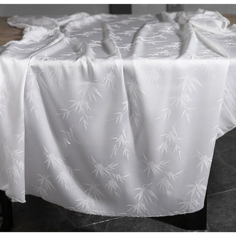 May include: A white tablecloth with a subtle bamboo leaf pattern. The fabric drapes over a table, with soft folds and shadows. The design features repeating white bamboo leaves on a white background, creating a delicate and elegant look.
