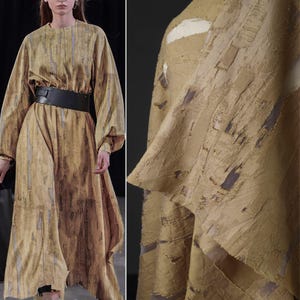 Op de afbeelding: Een lange, vloeiende jurk in tinten bruin en beige met verticale streepmotieven. De jurk heeft lange mouwen, een brede zwarte riem en een unieke, getextureerde stof. De afbeelding toont ook een close-up van de details van de stof.