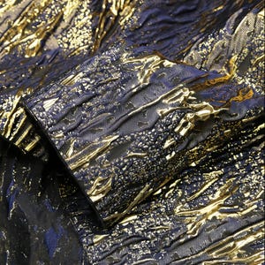 May include: Close-up of a textured fabric with a metallic gold and navy blue design. The fabric has a crinkled appearance with raised gold accents, creating a luxurious and shimmering effect. The fabric appears to be a high-end textile.