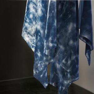 May include: Several flowing, sheer fabrics hang from a rod. The fabrics feature a tie-dye pattern in shades of indigo and white, creating a cloudy, marbled effect. The textiles appear lightweight and airy, suggesting they are scarves or shawls.